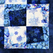 A close-up of a quilted fabric panel with a patchwork design in shades of blue, navy, and cream. The pattern features floral prints, swirling floral embroidery, and solid dark blue sections, framed by a dark blue border.