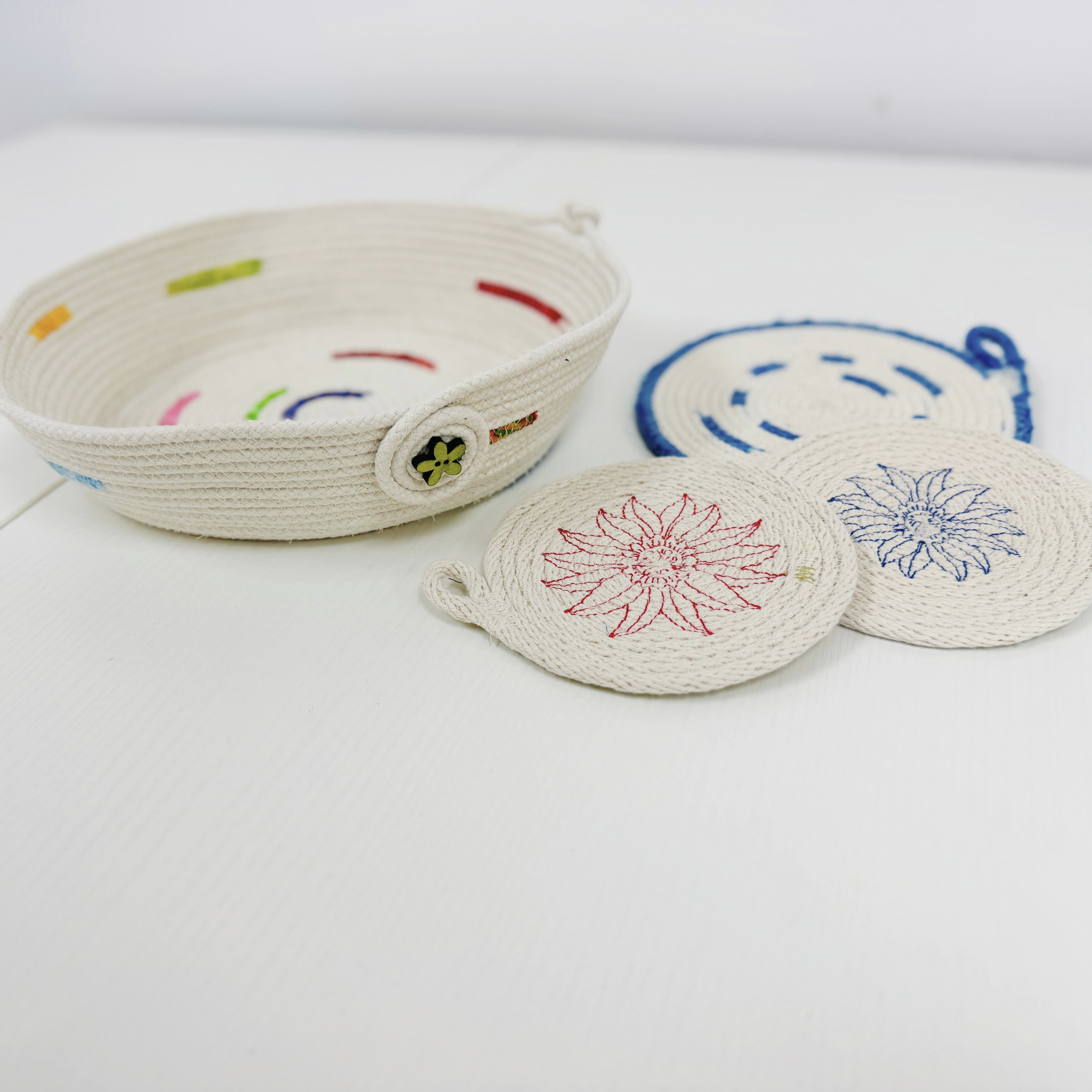 A woven rope basket with colorful embroidered accents sits on a white surface next to two matching round woven coasters with floral patterns in red and blue.