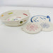 A woven rope basket with colorful embroidered accents sits on a white surface next to two matching round woven coasters with floral patterns in red and blue.
