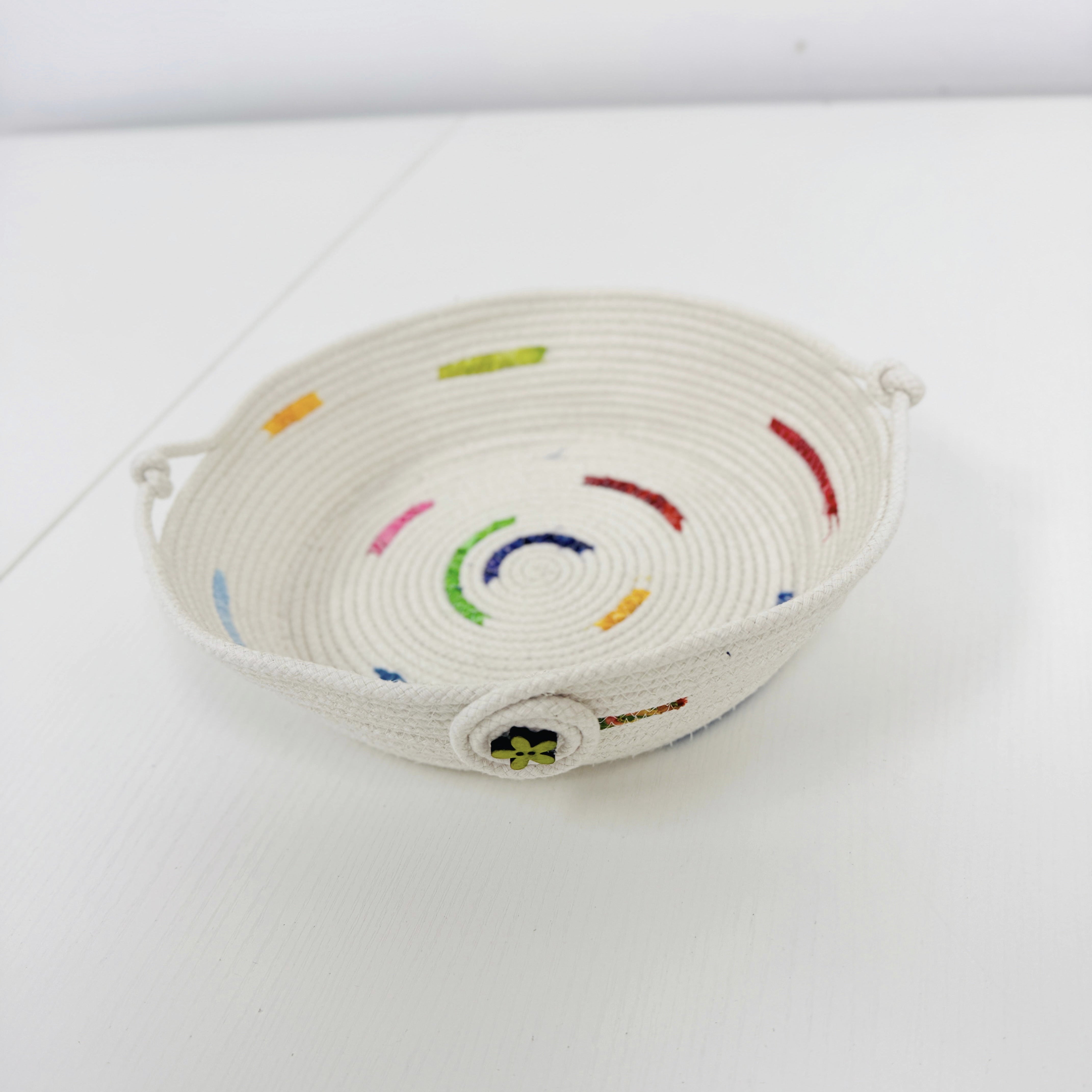 A small woven basket tray made from off-white rope, decorated with colorful embroidered curved lines and small dots in red, green, blue, yellow, and pink, set against a plain white background.