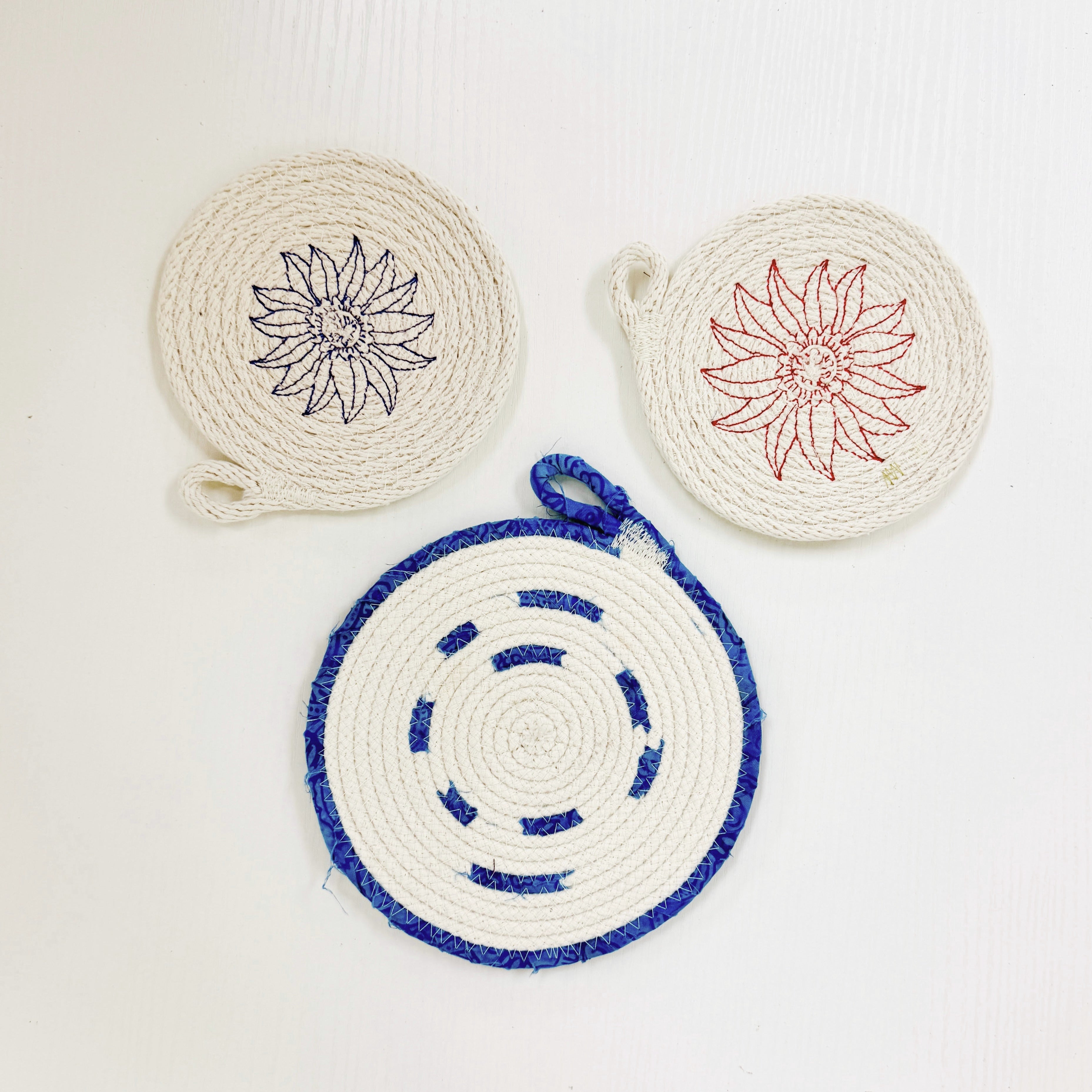 Three round woven rope coasters or trivets on a white background, featuring floral line-art designs in black, red, and blue.